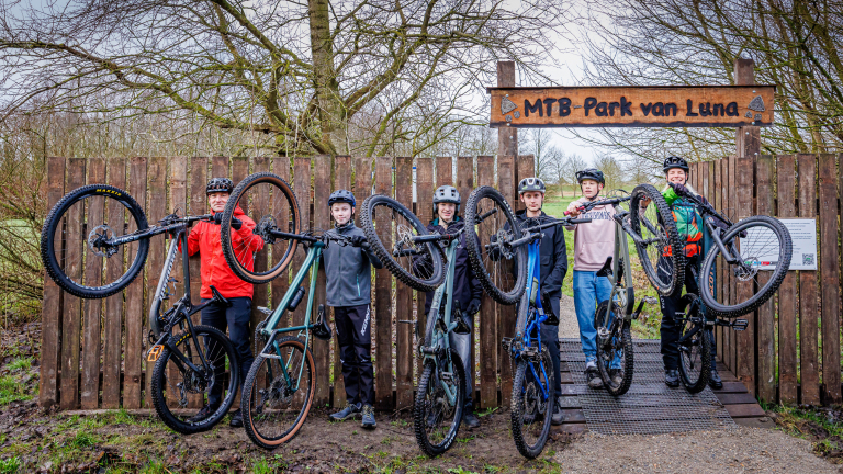 Een groep mountainbikers tilt hun fietsen op bij de ingang van MTB-Park van Luna, omgeven door bomen.