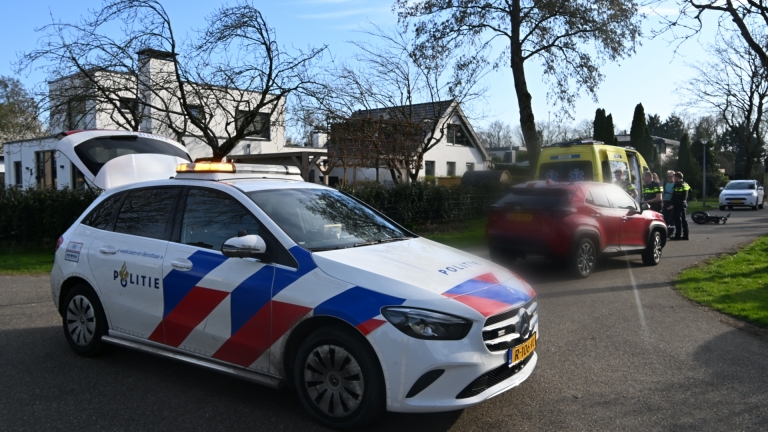 Een ambulance staat geparkeerd naast een rode auto op een straat, terwijl twee mensen zich buigen over spullen op de grond.