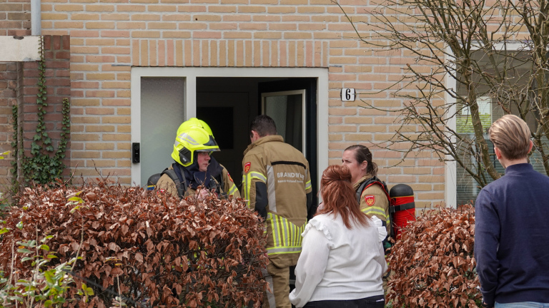 Brandweerlieden bij een rijtjeshuis, terwijl omstanders en geparkeerde auto's op straat zichtbaar zijn.