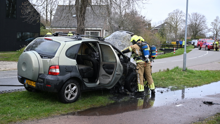 Brandweerlieden blussen een uitgebrande auto langs de weg met een brandweerwagen op de achtergrond.