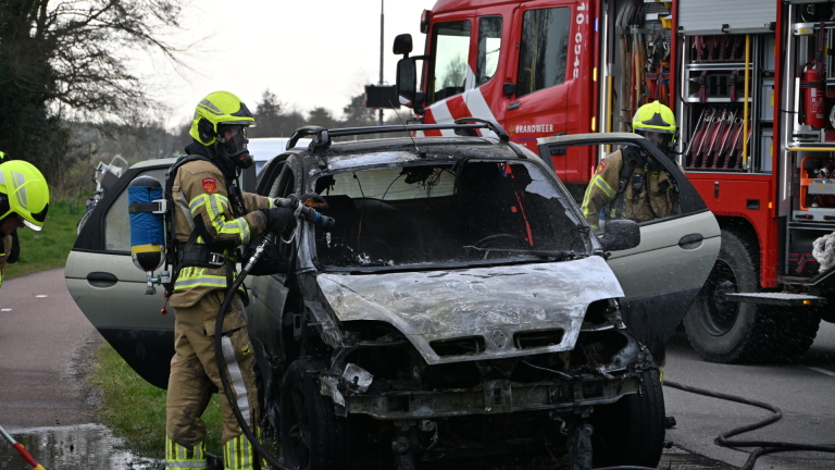 Brandweerlieden blussen een uitgebrande auto langs de weg met een brandweerwagen op de achtergrond.