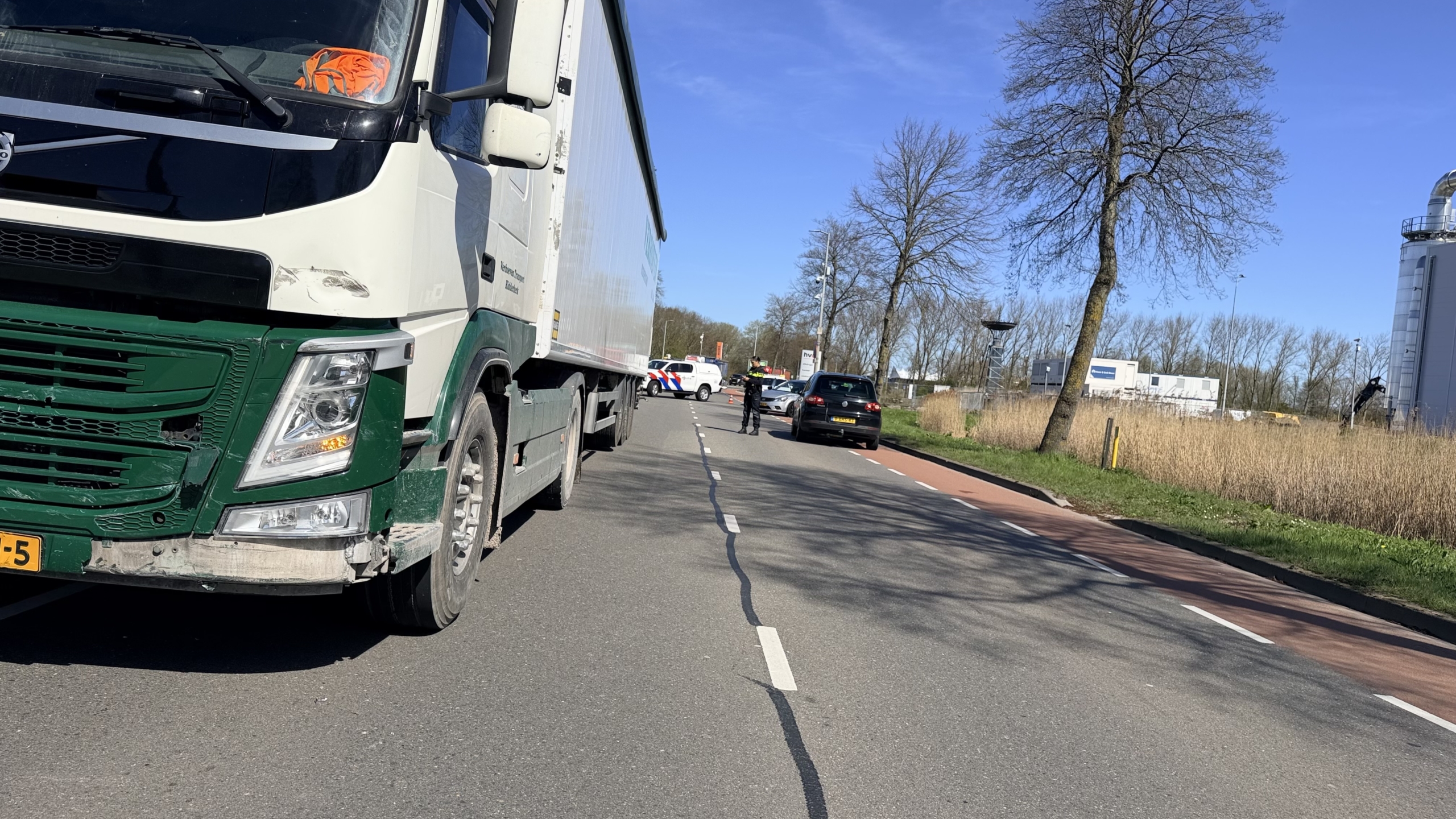Een zilveren auto met zware zijkantschade staat naast de weg; een politieagent en een persoon in oranje veiligheidskleding zijn erbij aanwezig.