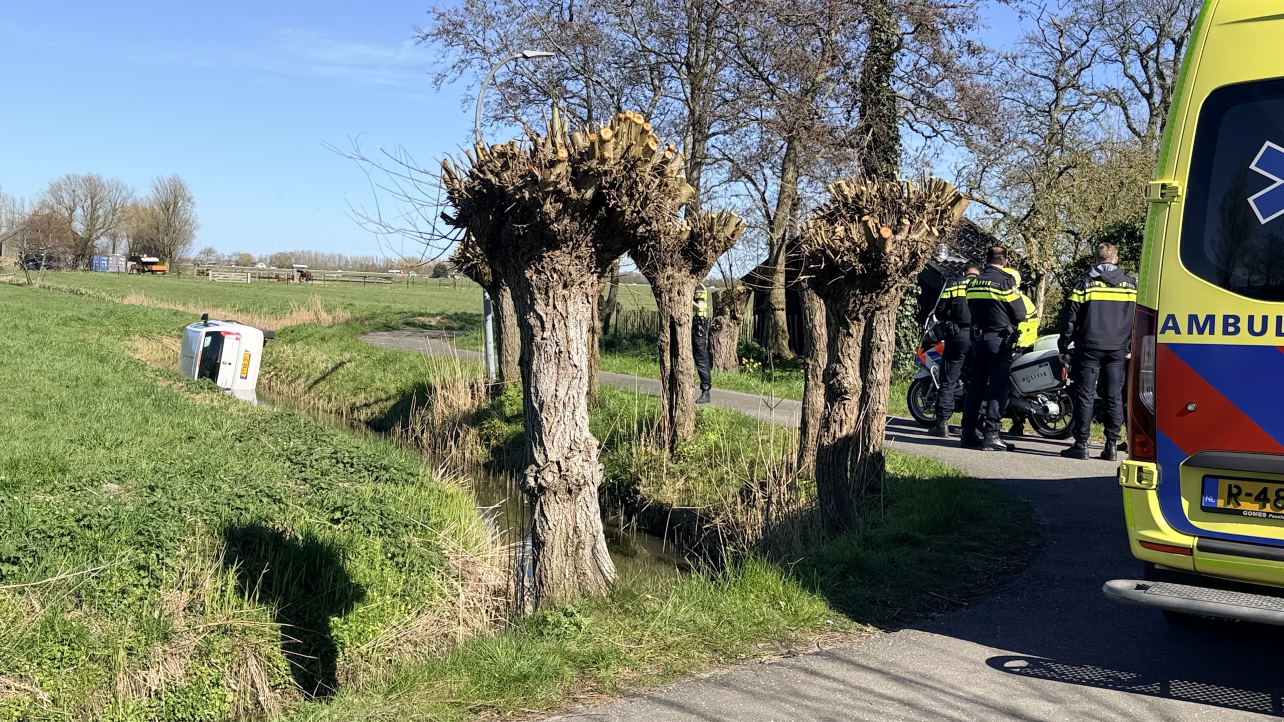 Auto op zijn kant in een sloot naast een grasveld en een weg in een landelijke omgeving.