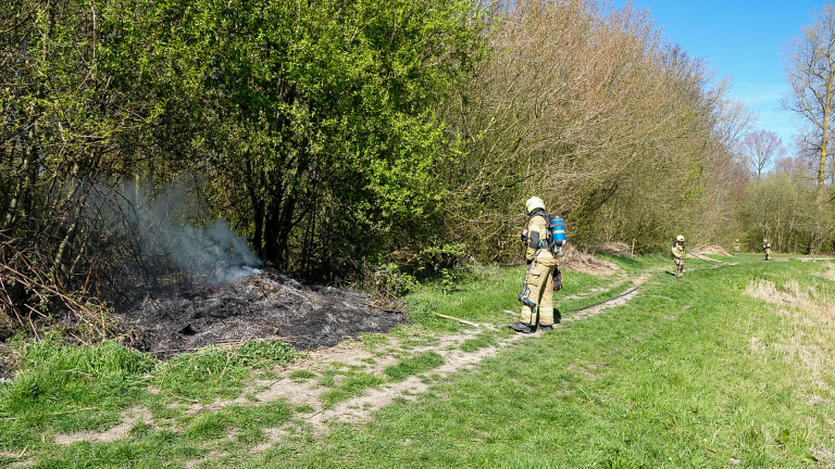 Brandweerman in beschermende kleding blust een smeulend stuk grond naast struiken.
