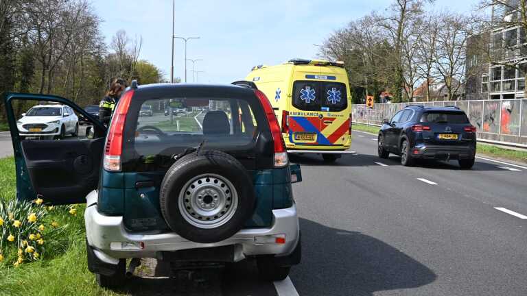 Een zwarte hatchback met een kapotte motorkap staat langs de weg, met politie en verkeer op de achtergrond.