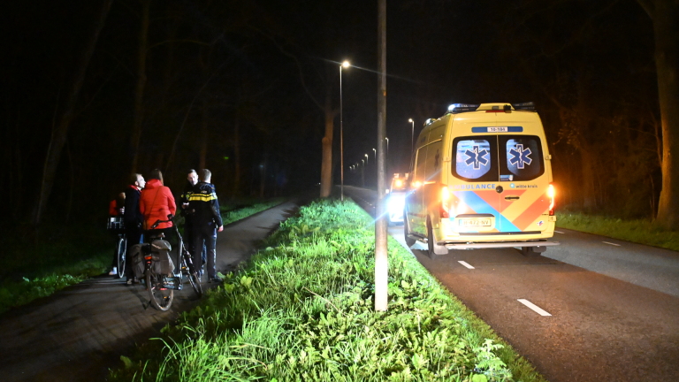 Een ambulance en een politieauto met zwaailichten staan 's nachts langs een weg, omgeven door mensen.