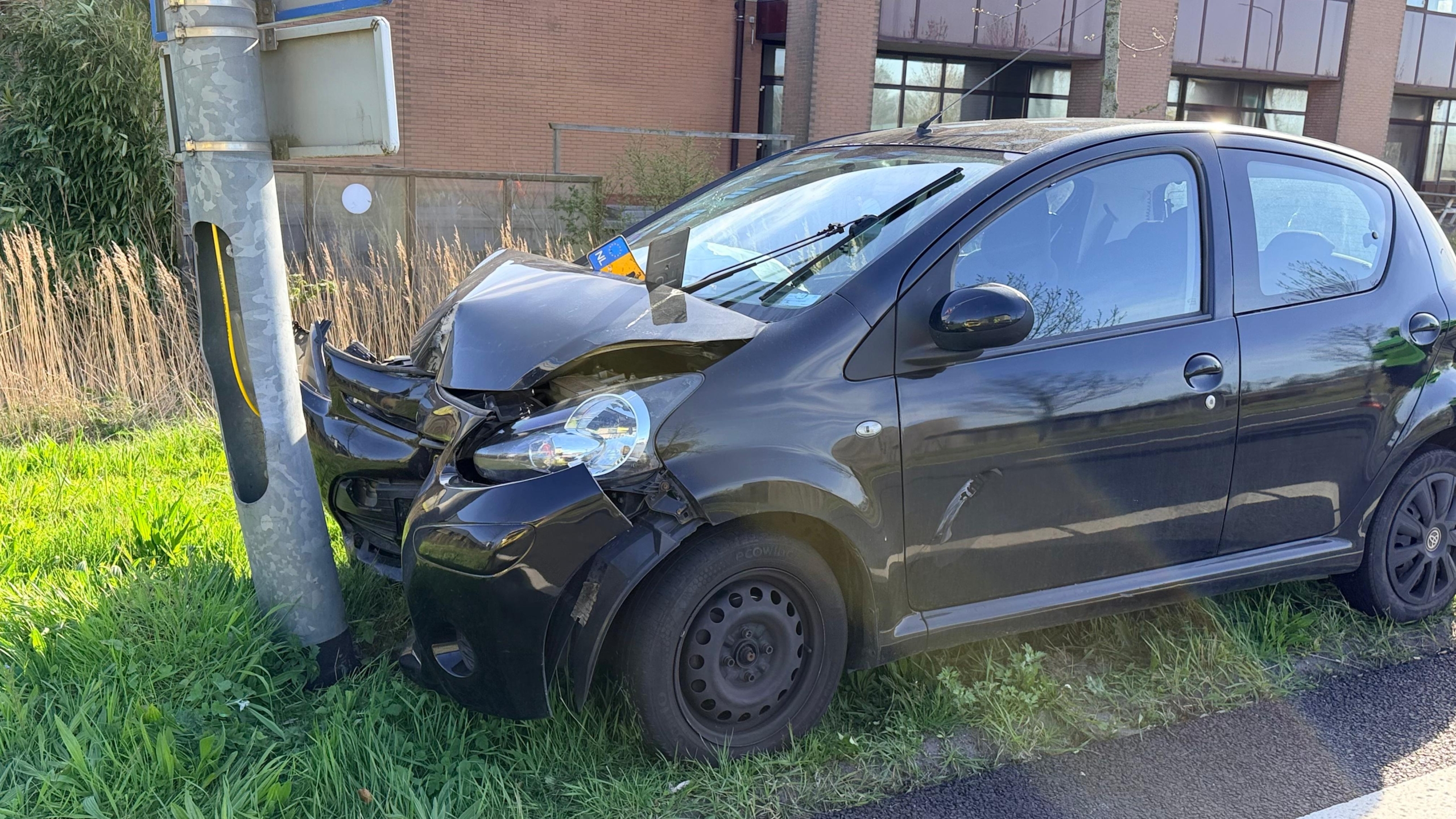 Auto met schade na botsing tegen een paal langs een weg, met verkeerskegel en mensen op de achtergrond.