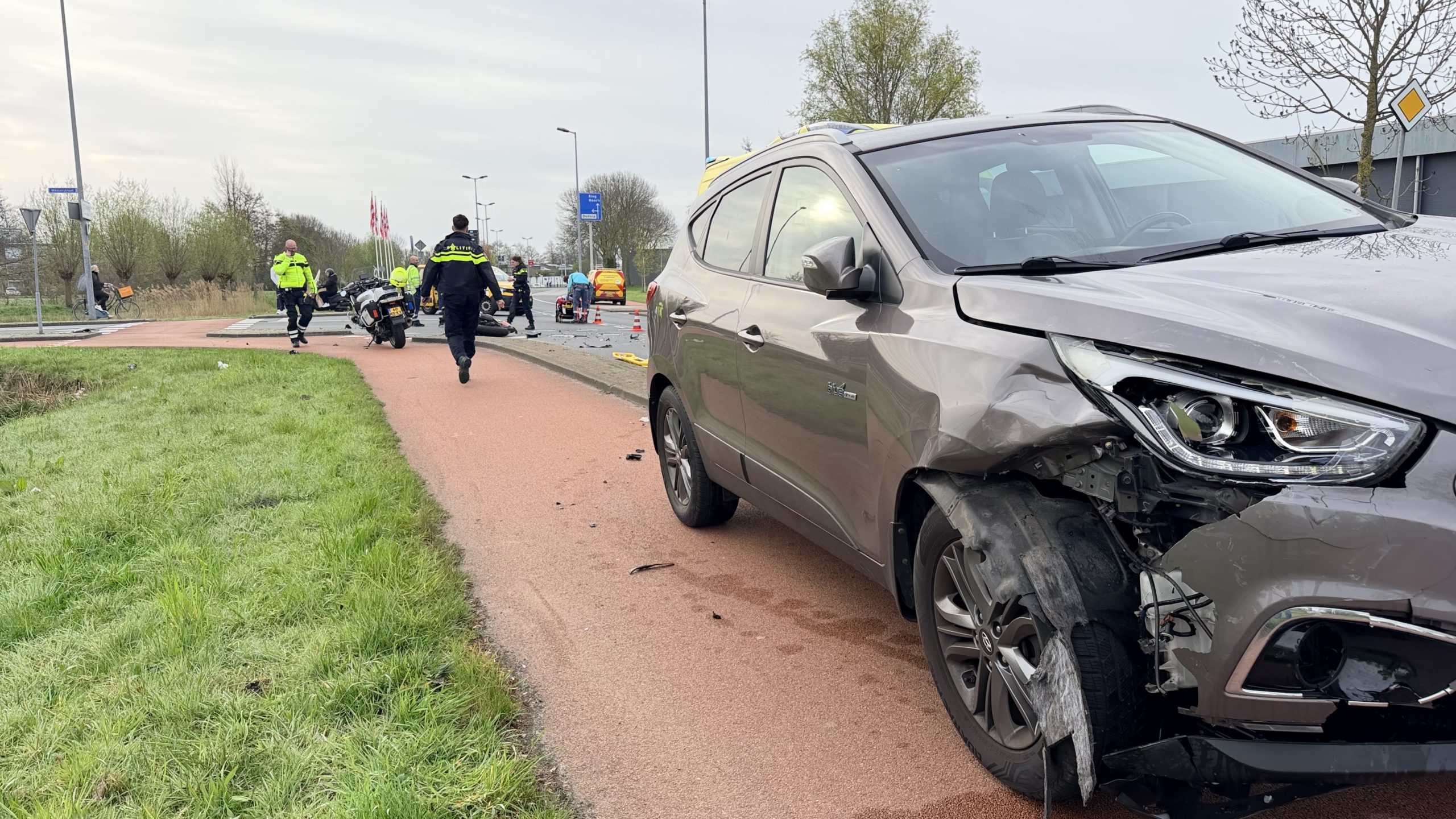 Verkeersongeluk met een omgevallen motorfiets, schade op de weg, en hulpverleningsvoertuigen, waaronder een ambulance en politiefiets.