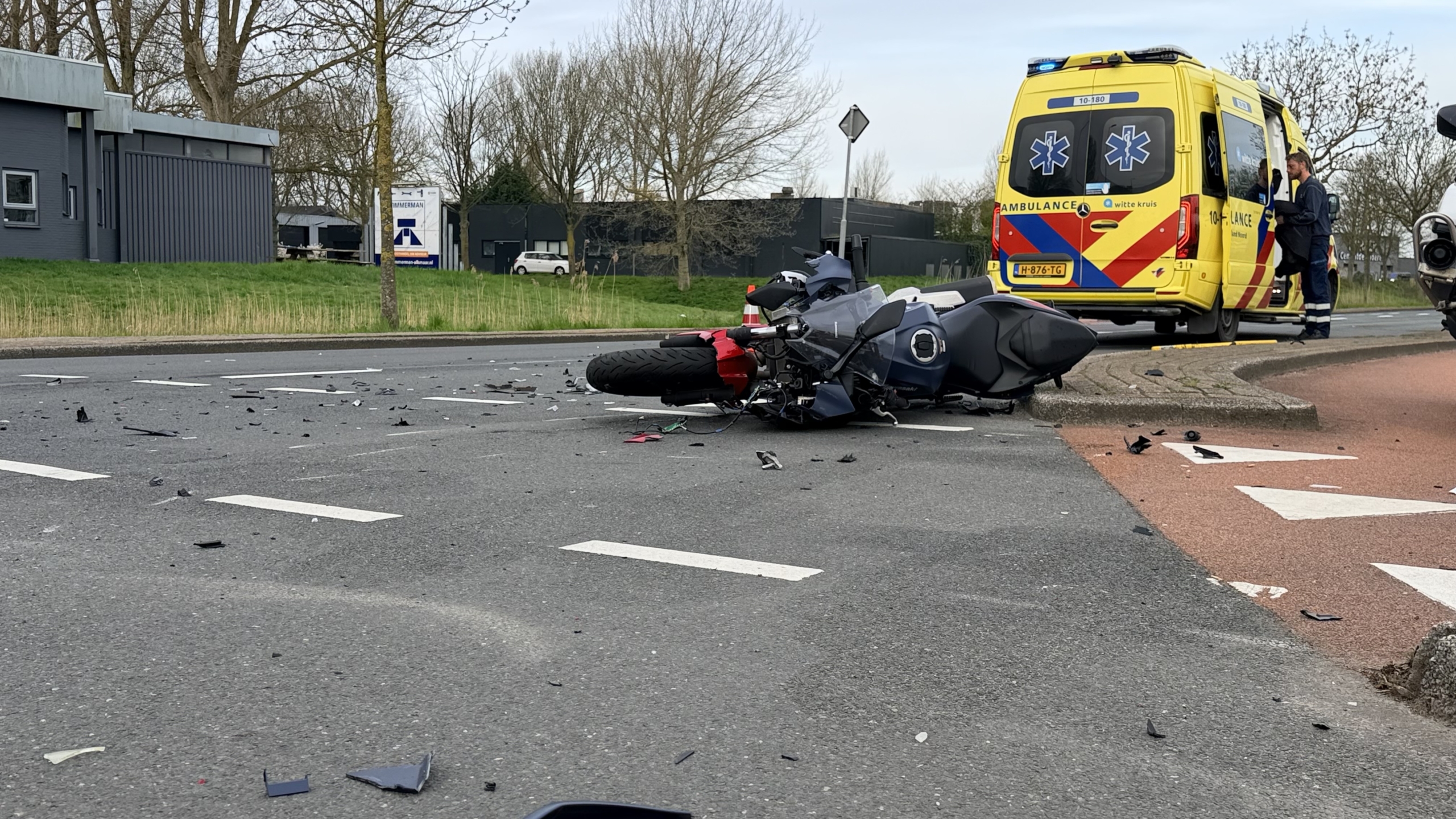 Verkeersongeluk met een omgevallen motorfiets, schade op de weg, en hulpverleningsvoertuigen, waaronder een ambulance en politiefiets.