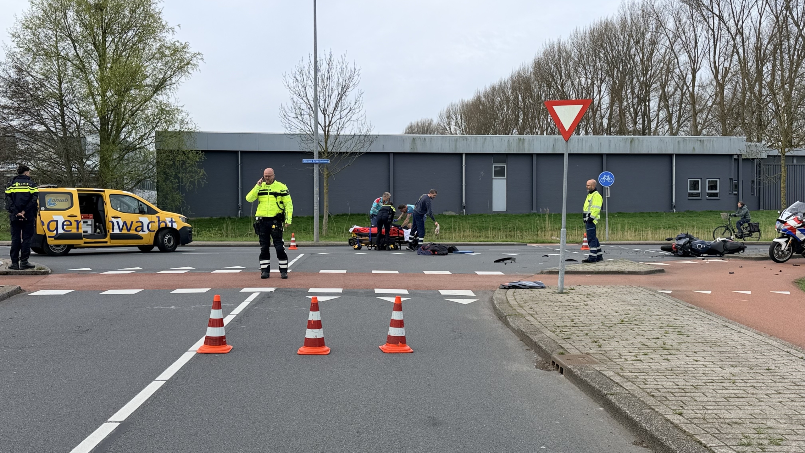 Verkeersongeluk met een omgevallen motorfiets, schade op de weg, en hulpverleningsvoertuigen, waaronder een ambulance en politiefiets.