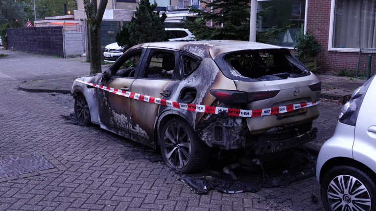Uitgebrande auto met geblakerd exterieur en politieafzetlint, geparkeerd op een straat.