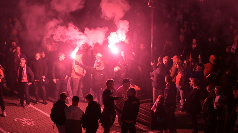 Twee mensen in gele jassen begeleiden een man op een plein, terwijl twee anderen op de achtergrond staan en kijken op hun telefoon.