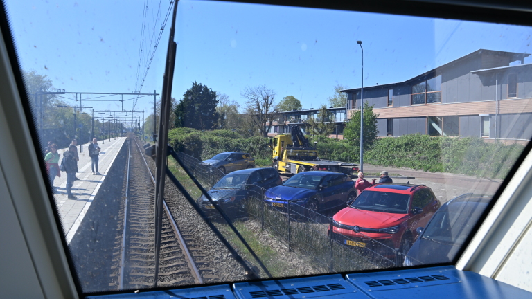 Auto tegen schuin hek met man in oranje vest die de deur opent. Politiewagen en omstanders op de achtergrond.
