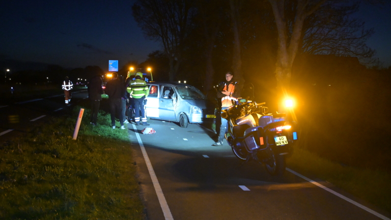 Auto met schade aan een kant van de weg in het donker, met een persoon en politie in de buurt.
