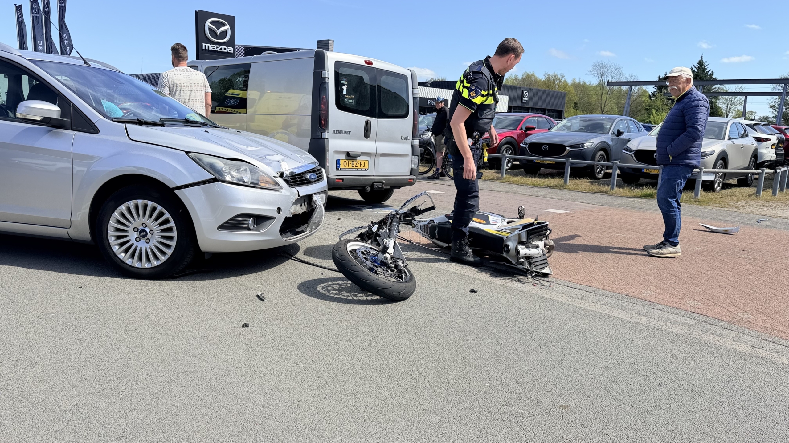 Politieagent onderzoekt een ongeval tussen een motorfiets en een grijze auto, nabij een showroom.