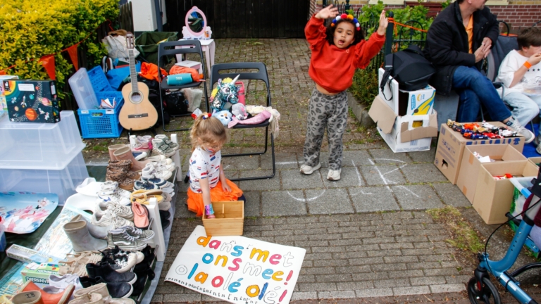 Mensen lopen langs een drukke markt met kraampjes en een Nederlandse vlag, nabij een historische kerk en gebouwen.