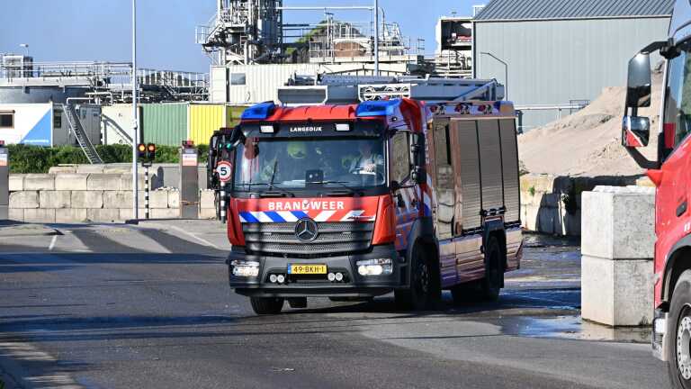 Brandweerwagens bij een industriële locatie met rook op de achtergrond en een groene windmolen aan de zijkant.