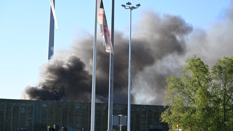 Brandweerwagens bij een industriële locatie met rook op de achtergrond en een groene windmolen aan de zijkant.