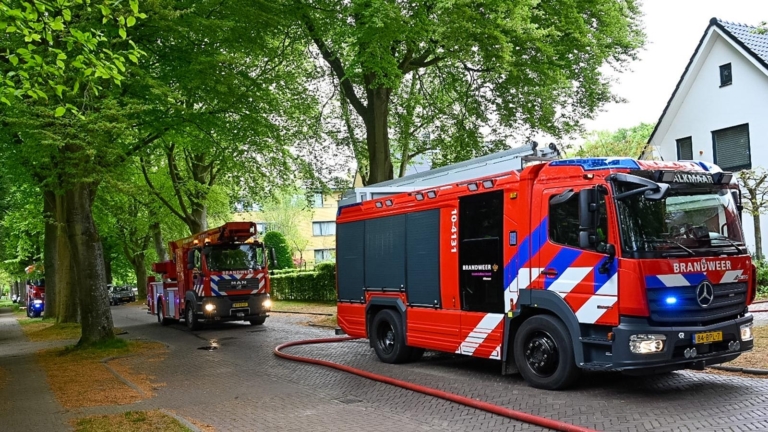 Wit huis met zwarte kozijnen, waar rook uitkomt bij een dakraam; rode bouwsteiger en afvalcontainer op de voorgrond.