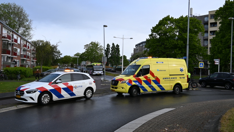Politieauto en ambulance staan geparkeerd langs een weg in een stedelijke omgeving met huizen en een bus op de achtergrond.