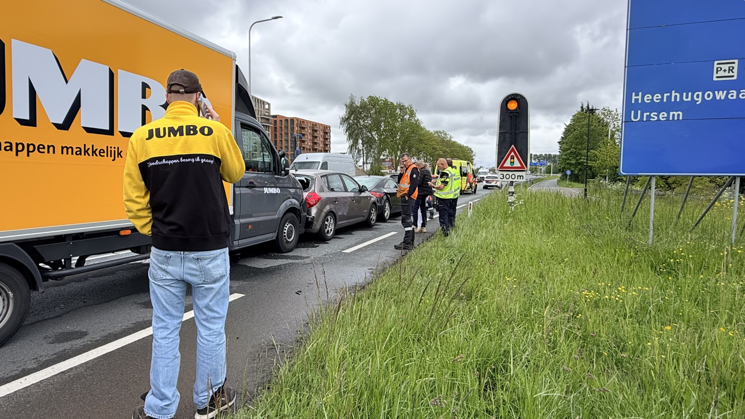 Een gele ambulance en een vrachtwagen zijn gestopt langs de weg, met mensen in veiligheidshesjes verderop.