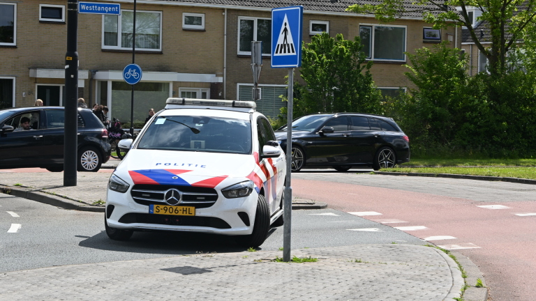 Fiets met tassen geparkeerd bij een rotonde, terwijl een politieauto voorbijrijdt naast twee andere auto's.
