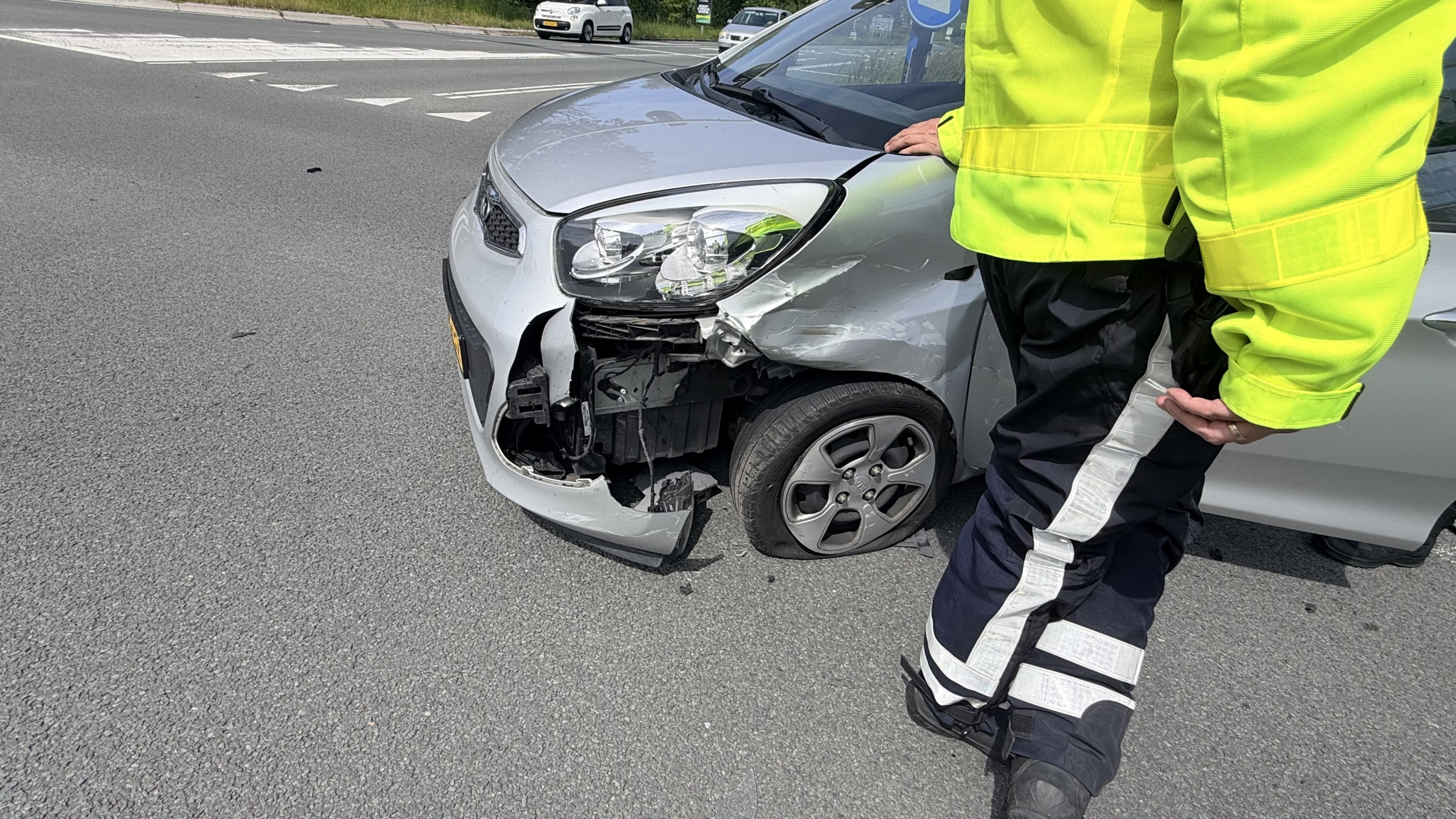 Een zwart voertuig met schade aan de voorkant, omringd door politieauto’s en -motorfiets, met agenten in gesprek op de achtergrond.