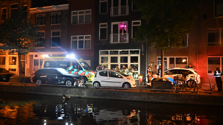 Nachtelijke scène met een ambulance en politieauto voor een gebouw, omringd door hulpverleners, met een persoon in het water.