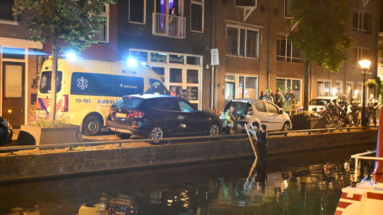 Nachtelijke scène met een ambulance en politieauto voor een gebouw, omringd door hulpverleners, met een persoon in het water.