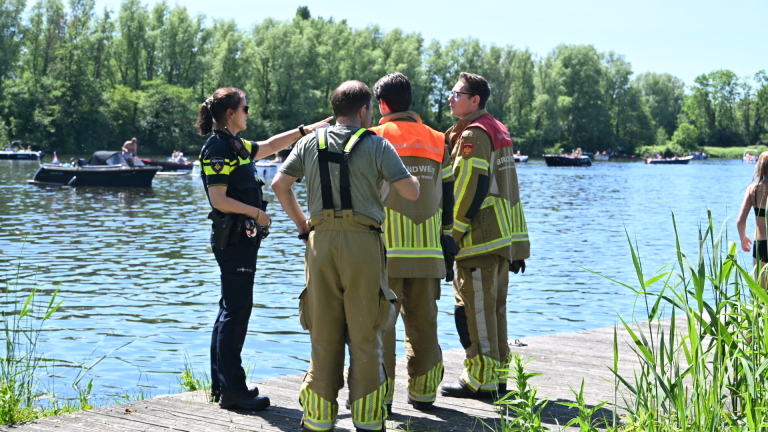 Politie- en brandweermensen staan op een steiger aan een meer, met boten en mensen in de achtergrond.