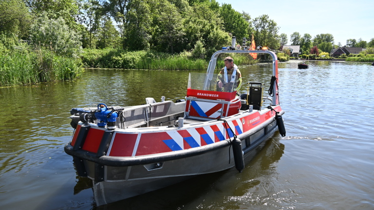 Politie- en brandweermensen staan op een steiger aan een meer, met boten en mensen in de achtergrond.