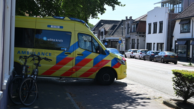Ambulancepersoneel helpt een persoon op de stoep naast een ambulance; een jongen kijkt toe.