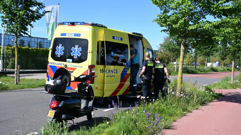 Ambulance en politie bij een incident langs de weg, met een politieagent en een scooter op de voorgrond.