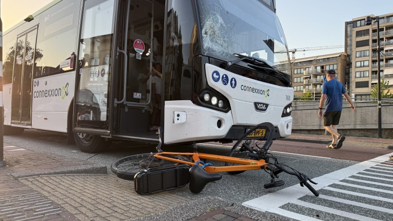 Een bus met een ingedeukte voorruit staat stil op straat met een oranje fiets eronder; een voorbijganger loopt langs.
