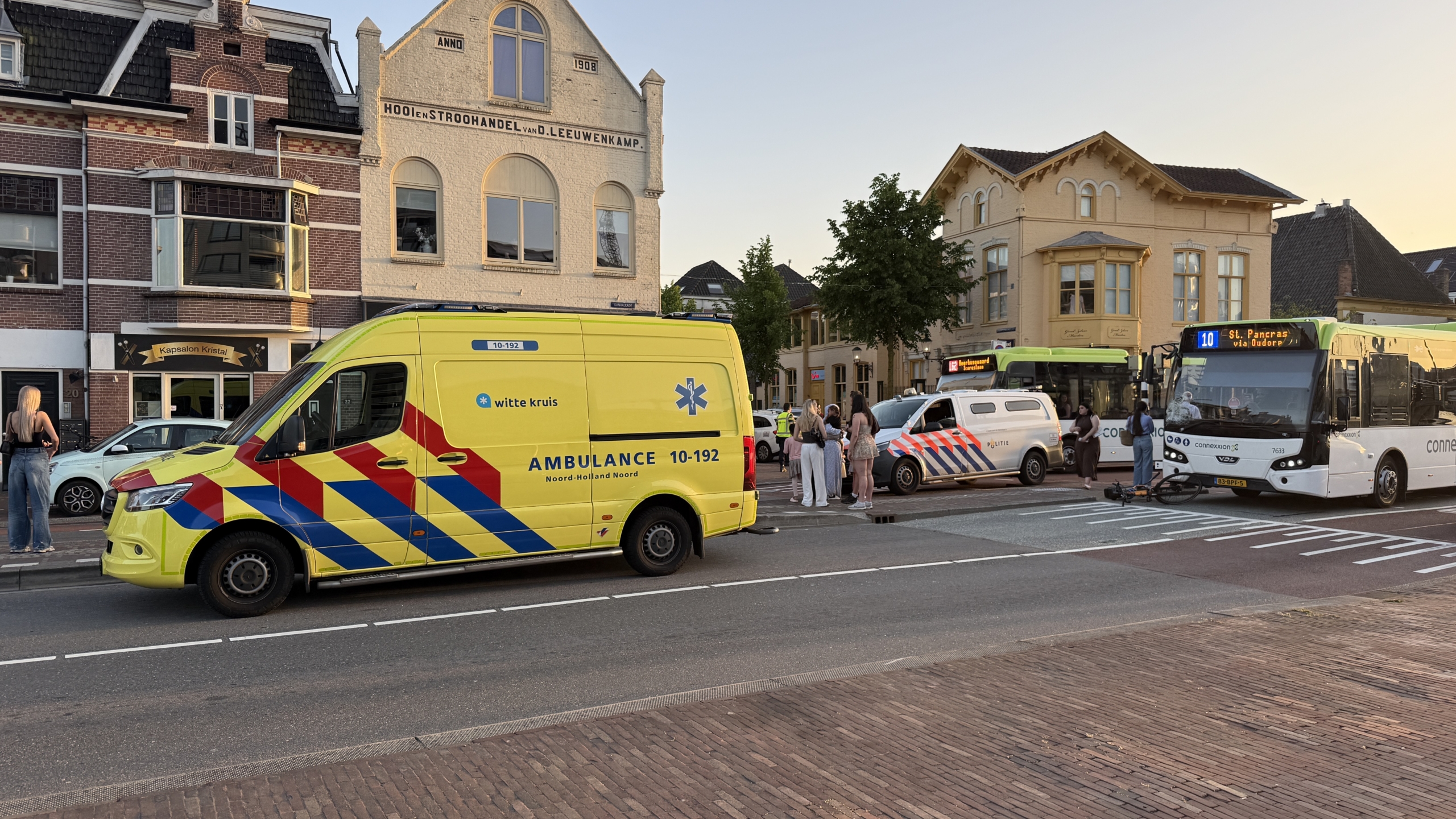 Een bus met een ingedeukte voorruit staat stil op straat met een oranje fiets eronder; een voorbijganger loopt langs.