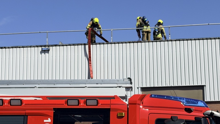 Brandweerlieden in gele helmen werken op een dak met een rode brandslang; een rode brandweerauto staat ernaast.
