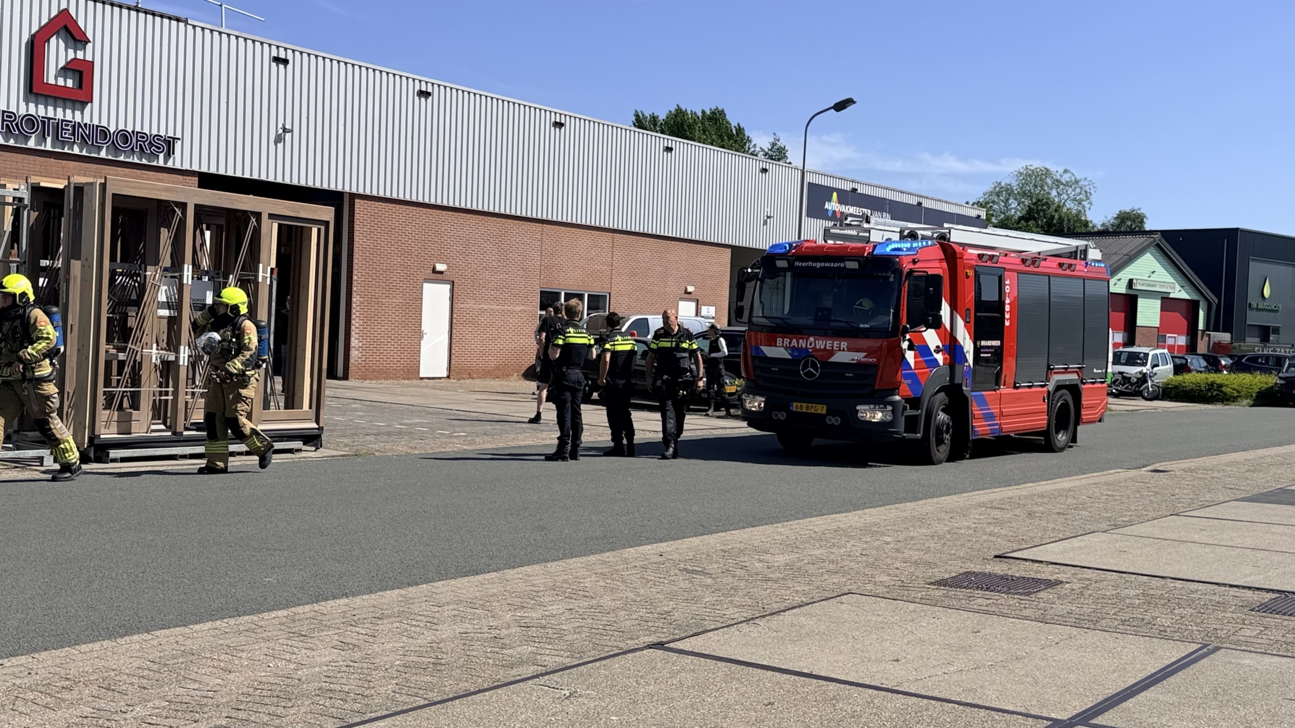 Brandweerlieden in gele helmen werken op een dak met een rode brandslang; een rode brandweerauto staat ernaast.