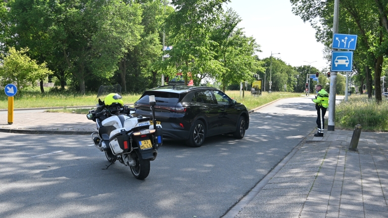 Een zwarte auto is botst met een grijze scooter op een straat; een ambulance staat op de achtergrond.