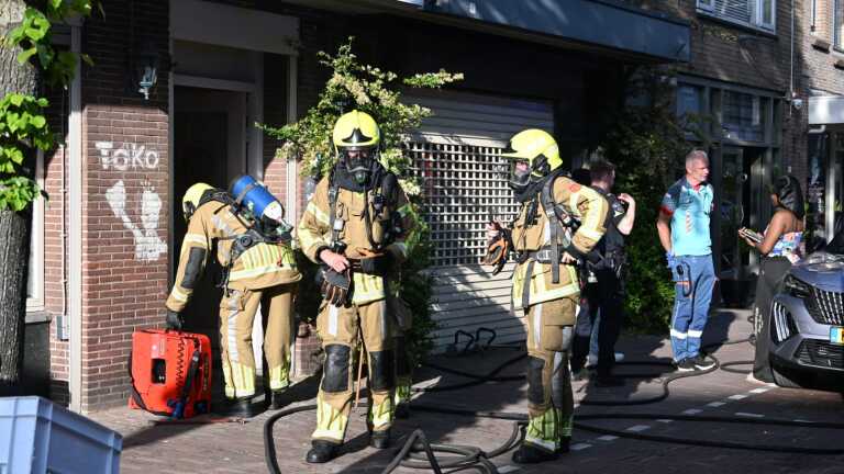Brandweermannen in gele helmen staan bij een brandweerwagen in een straat met winkels in een rijtje.