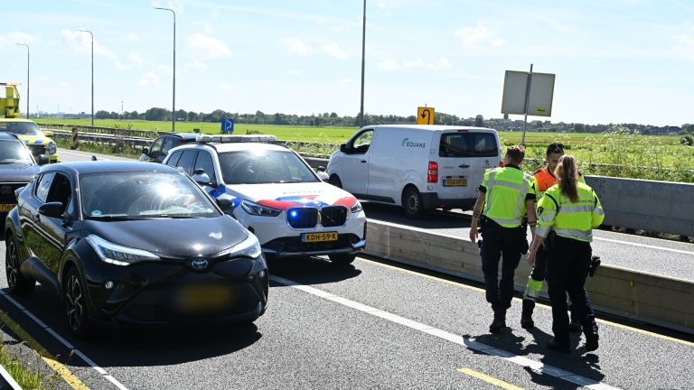 Een zwarte auto wordt door een gele sleepwagen opgeladen op een snelweg, terwijl een man in veiligheidskleding de weg veegt.
