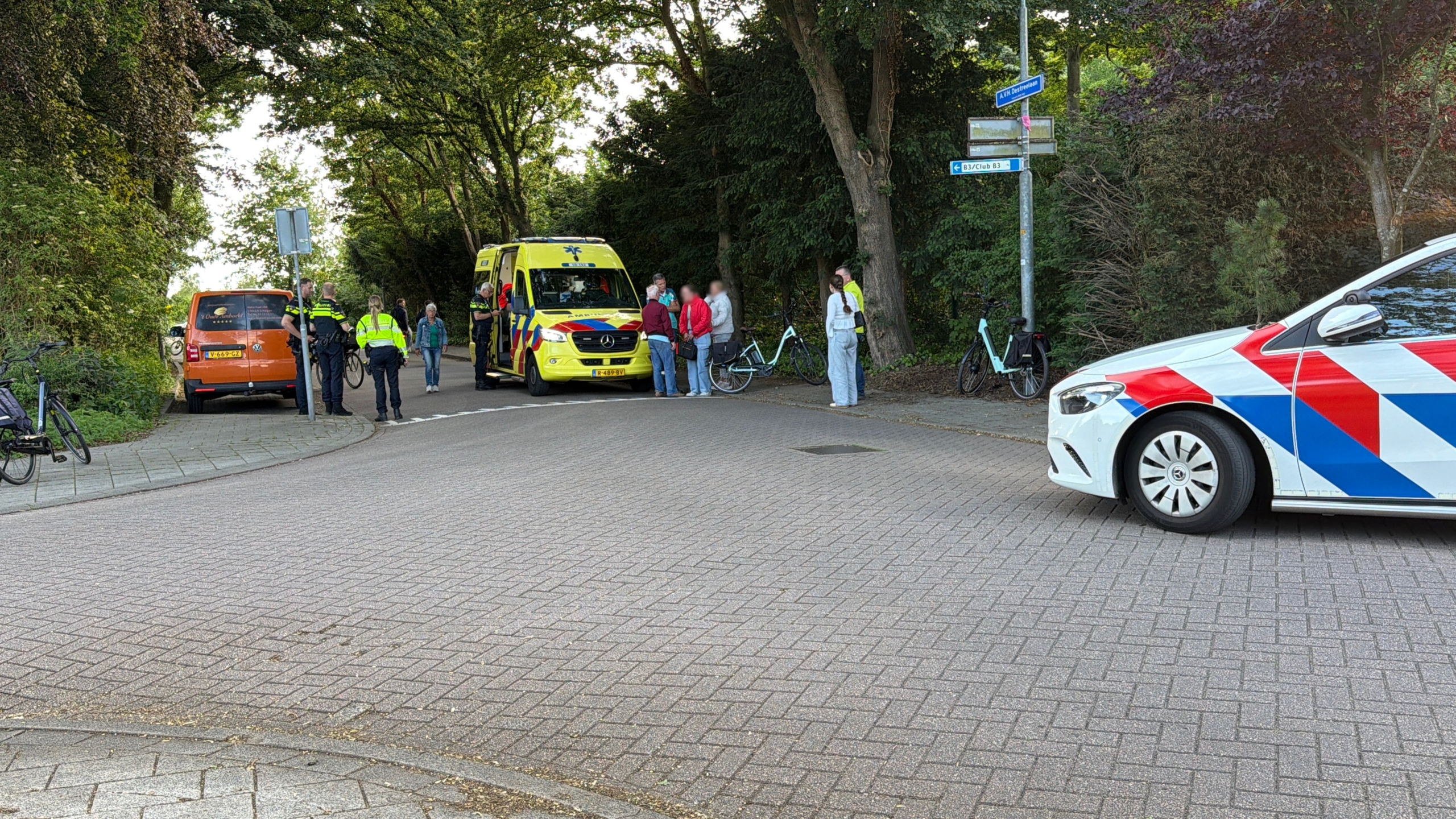 Twee politieagenten staan bij een geparkeerde ambulance, terwijl een vrouw verderop aan de telefoon is.