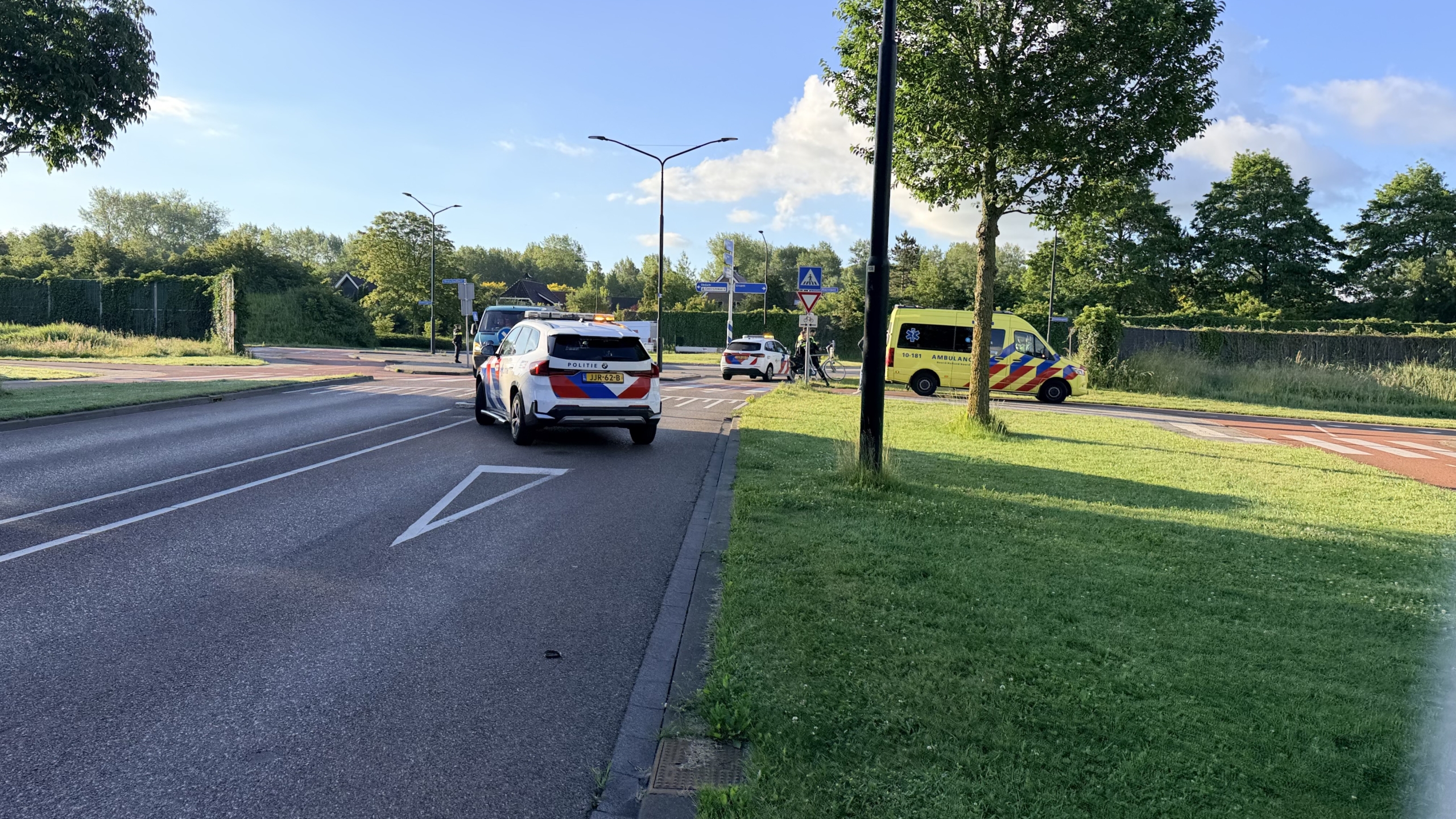 Een gele ambulance staat geparkeerd langs de weg bij een kruispunt, met politieagenten en een fiets in de buurt.