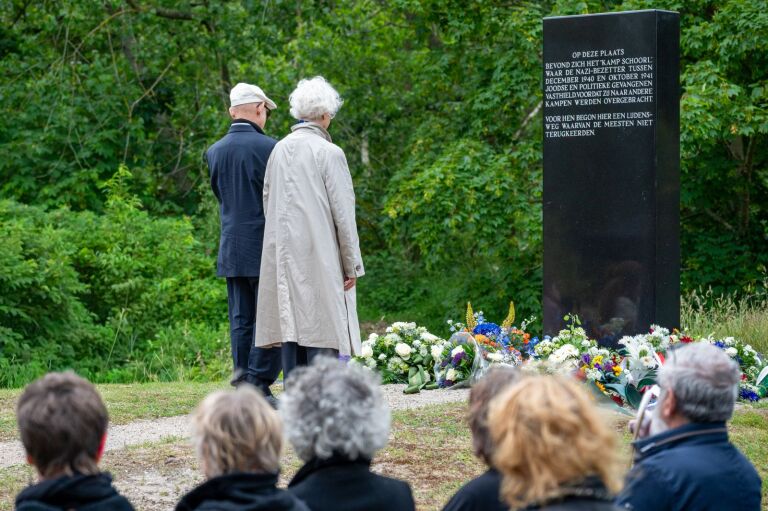 Twee mensen bekijken een herdenkingsmonument omgeven door bloemen, met toeschouwers op de voorgrond in een groene omgeving.