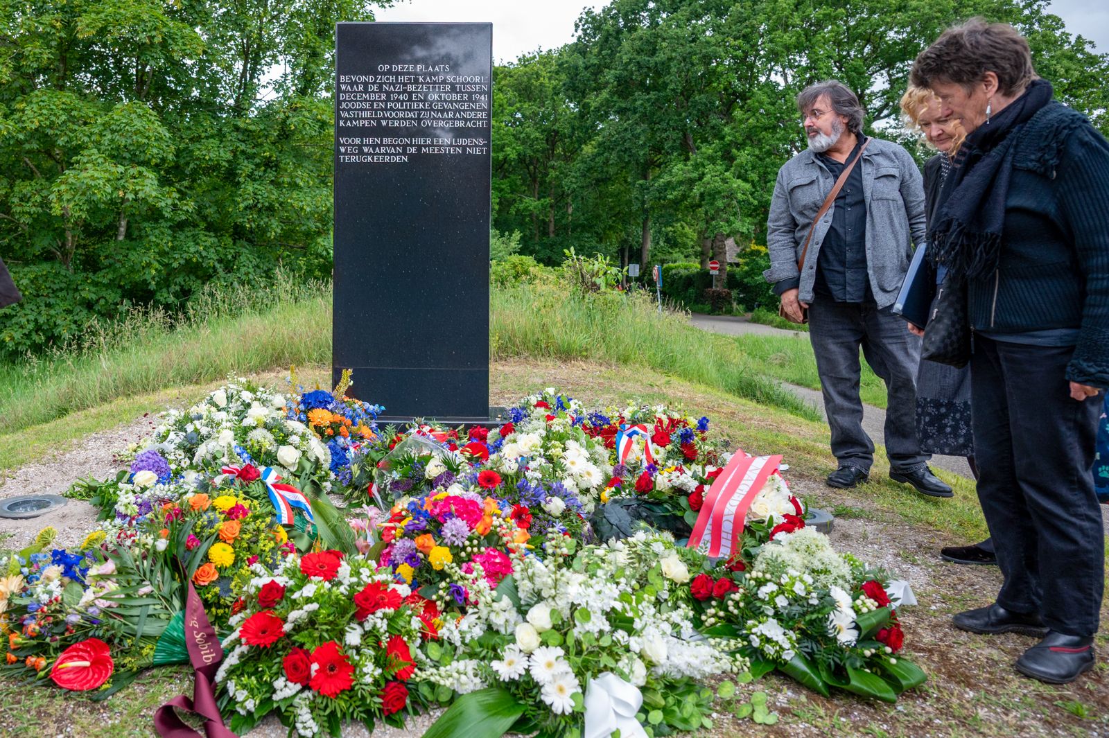 Twee mensen bekijken een herdenkingsmonument omgeven door bloemen, met toeschouwers op de voorgrond in een groene omgeving.