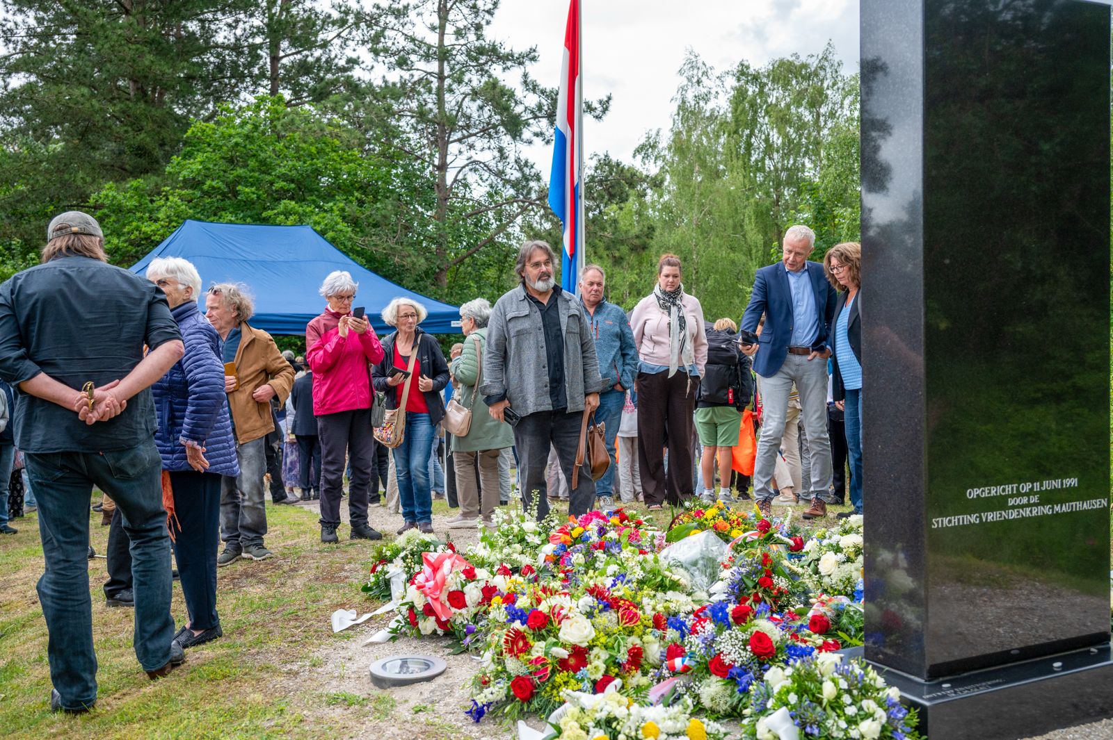 Twee mensen bekijken een herdenkingsmonument omgeven door bloemen, met toeschouwers op de voorgrond in een groene omgeving.