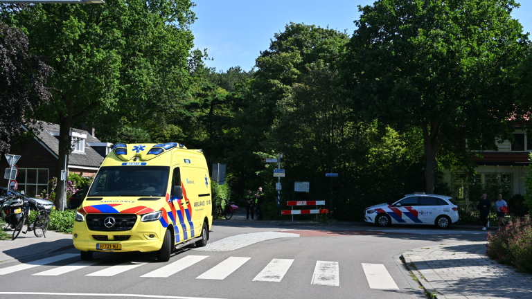 Politie en ambulance bij een incident met een zwarte auto op een zonnige dag, met een motoragent op de stoep.