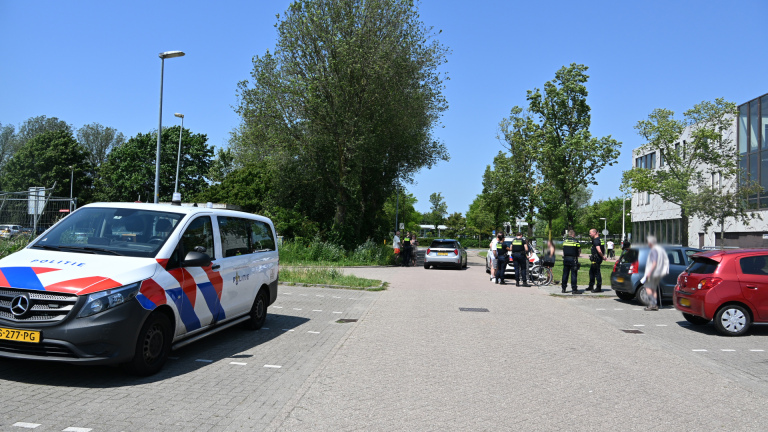 Politieauto en agenten op straat in gesprek met mensen, omgeven door bomen en een kantoorgebouw op de achtergrond.