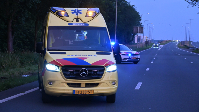 Zwaar beschadigde auto naast de weg in hoog gras, omgeven door bomen.
