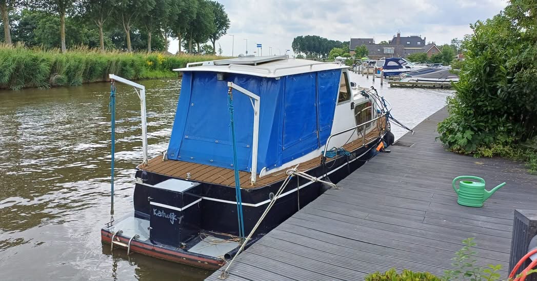 Blauw motorjacht aangemeerd aan een houten steiger bij een kanaal, omgeven door groen en bomen.