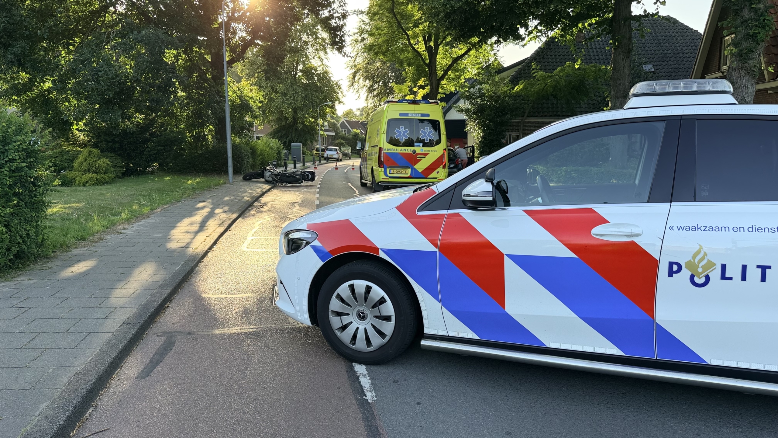 Omgevallen motorfiets op de stoep naast een ambulance en politieauto, met omstanders aan de kant van de weg.